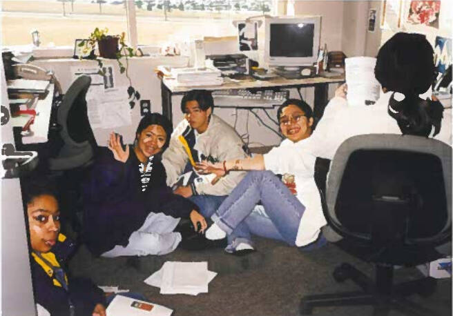 Students sitting on office floor holding papers smiling at camera